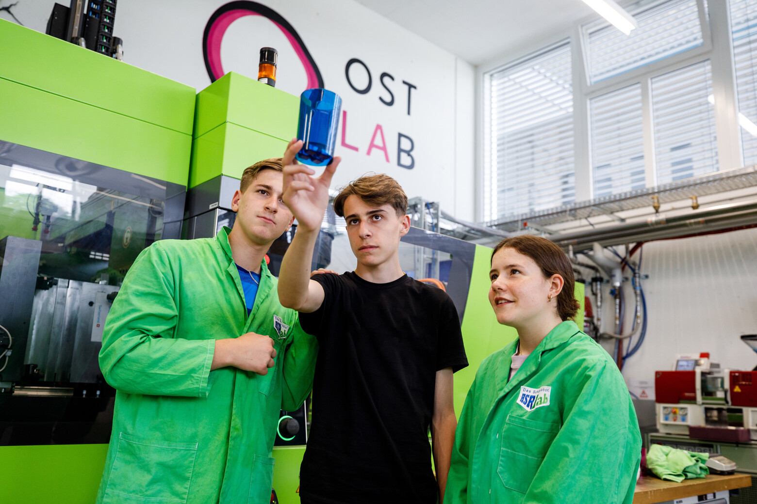 Zwei Schüler und eine Schülerin betrachten einen blauen, transparenten Kunststoffbecher, den sie im OSTLAB hergestellt haben.