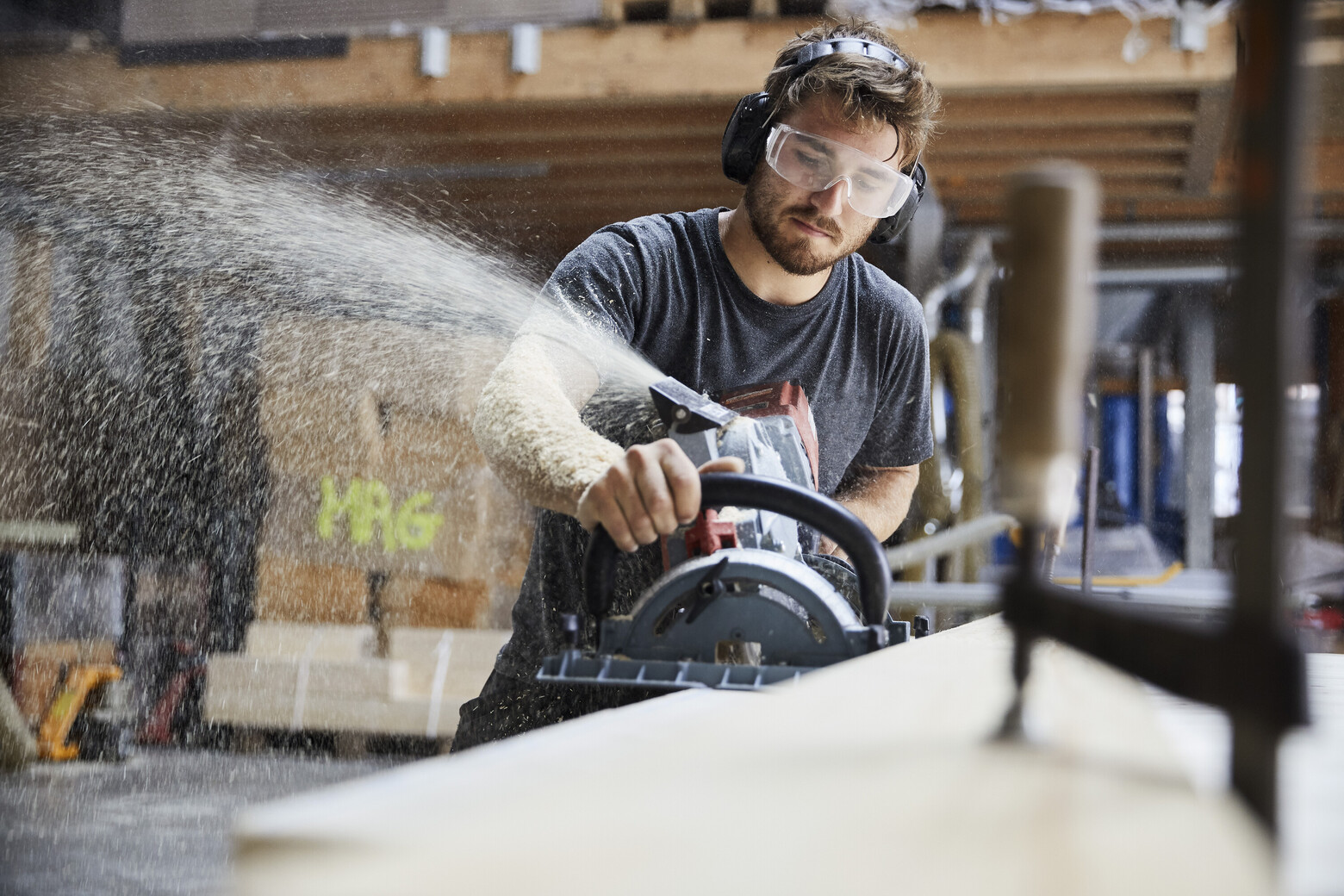 Ein Zimmermann fräst gerade das Holz und die Sphäne fliegen im gegen die Schutzbrille.