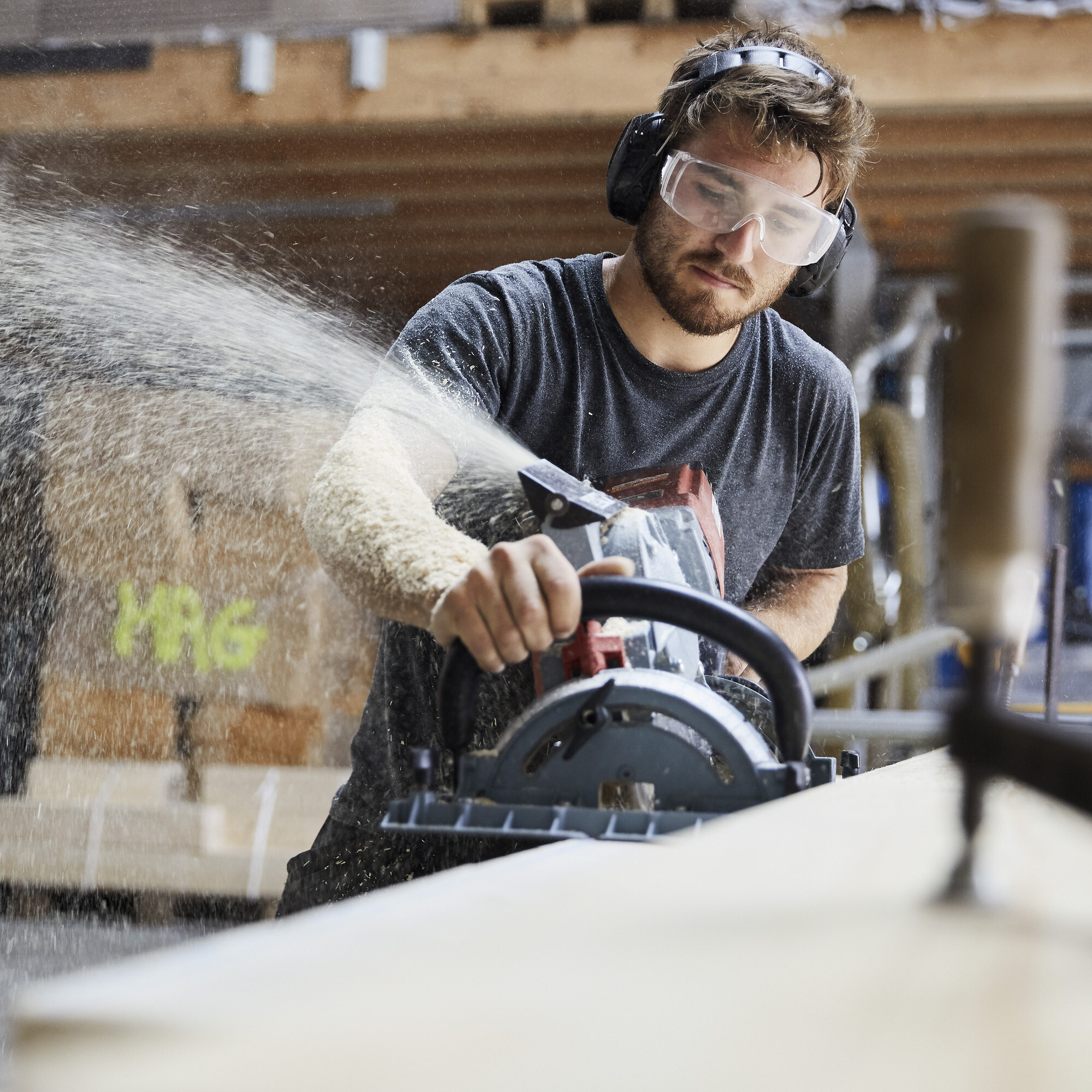 Ein Zimmermann fräst gerade das Holz und die Sphäne fliegen im gegen die Schutzbrille.
