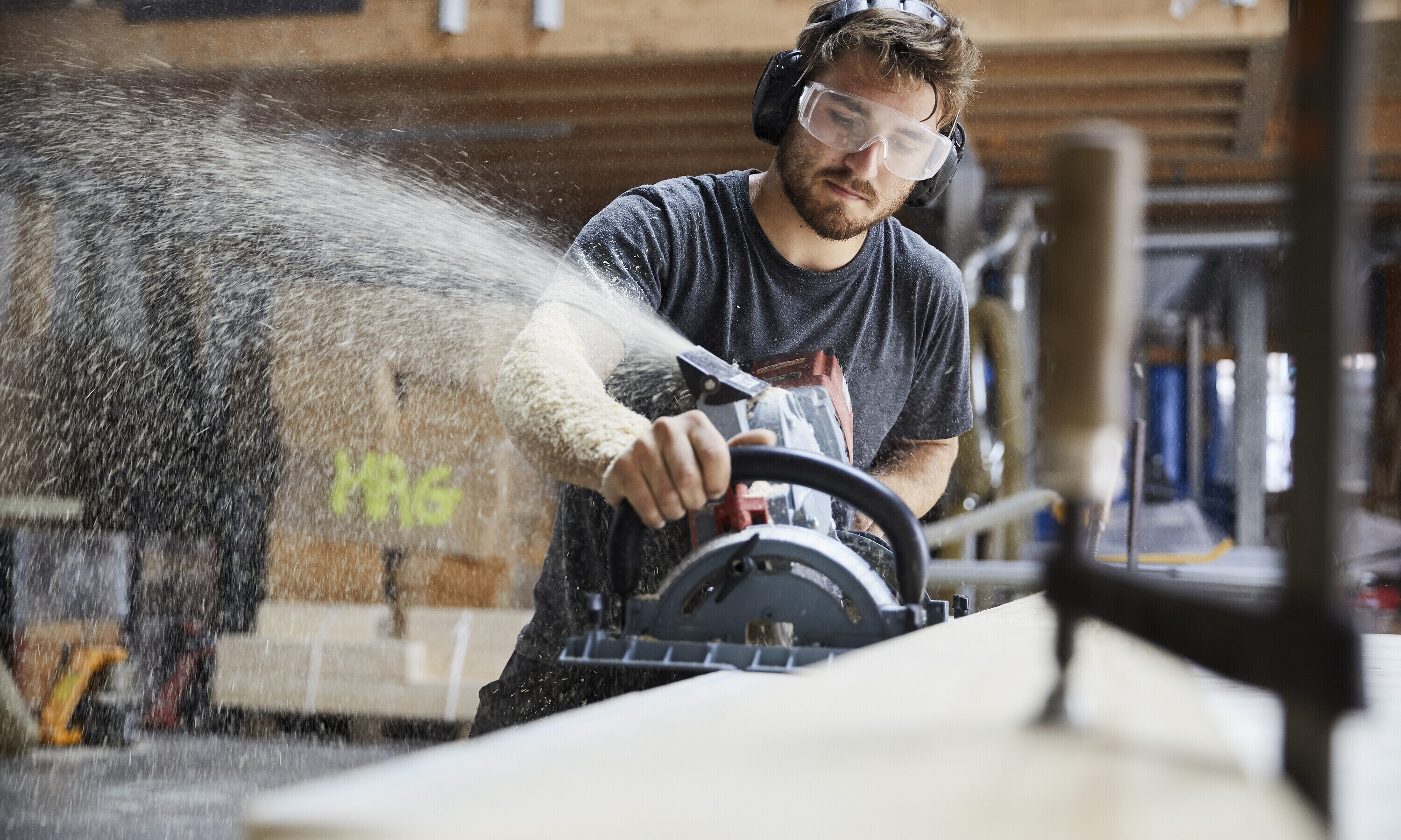 Ein Zimmermann fräst gerade das Holz und die Sphäne fliegen im gegen die Schutzbrille.