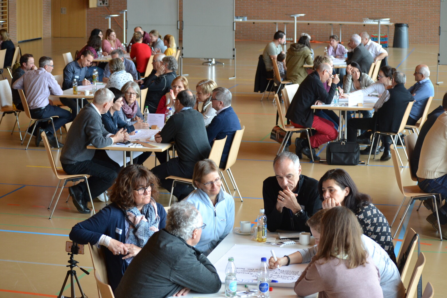 In einer Turnhalle sitzen mehrere Gruppen von Menschen an Tischen und besprechen ihre Themen.