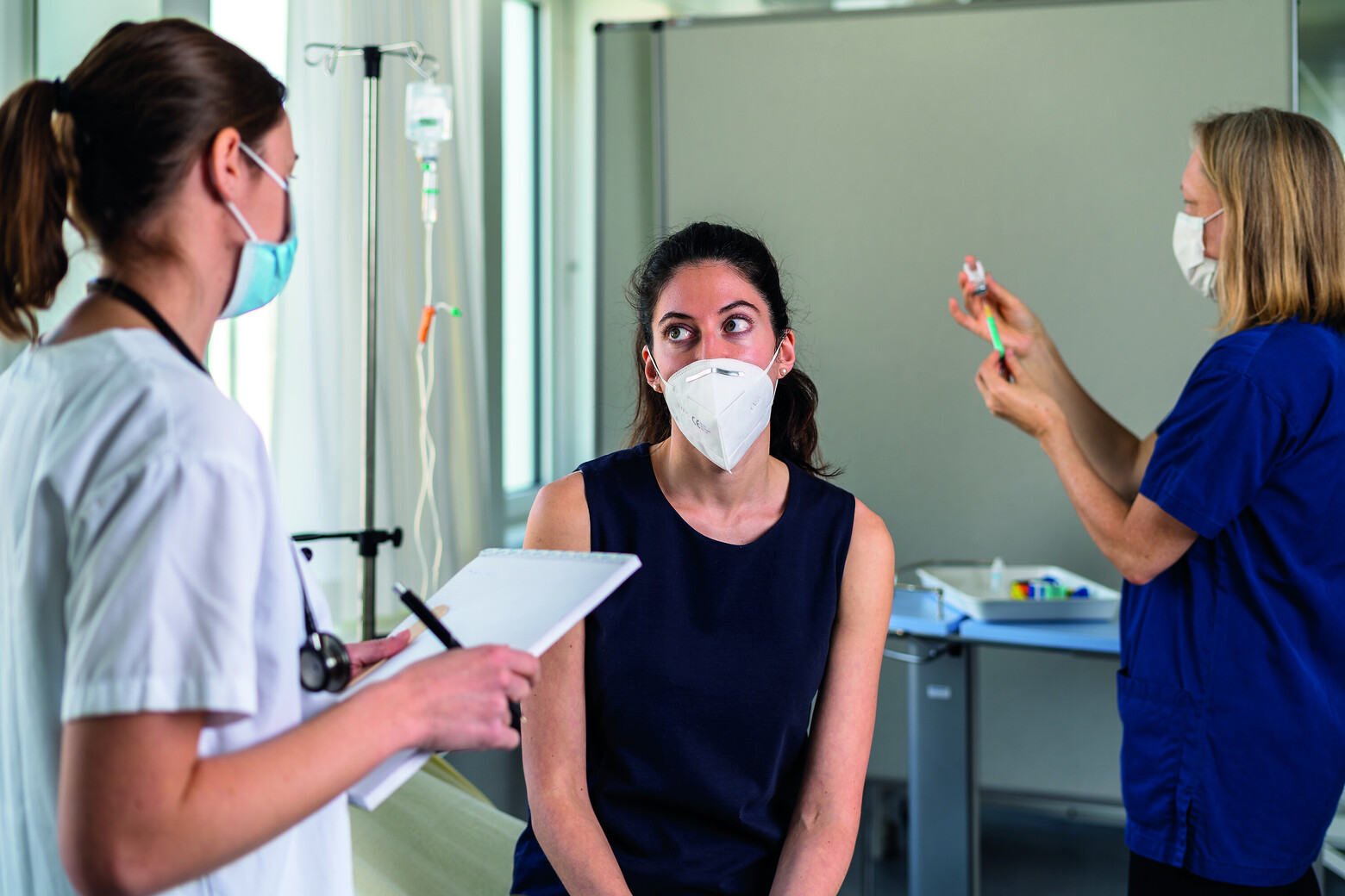 Eine Patientin mit FFP2-Maske sitzt auf dem Therapietisch, während eine Pflegefachfrau mit normalem Mundschutz sie befragt und behandelt. Im Hintergrund bereitet ein Teammitglied die Untersuchung vor.