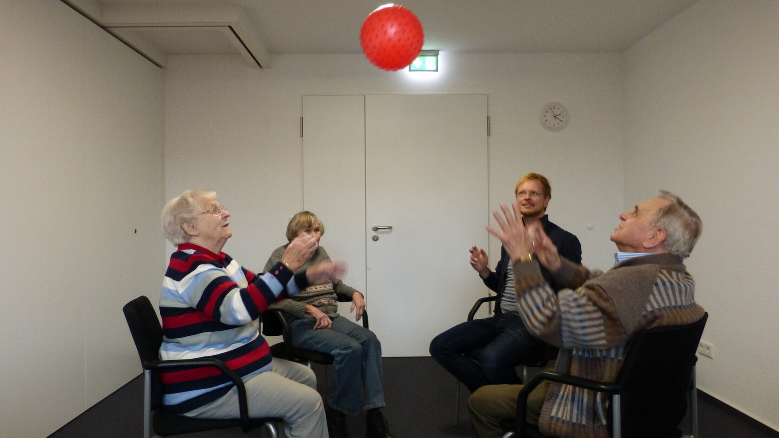 Vier Bewohnende sitzen mit den Stühlen im Kreis und werfen einen roten Luftballon in die Höhe. 