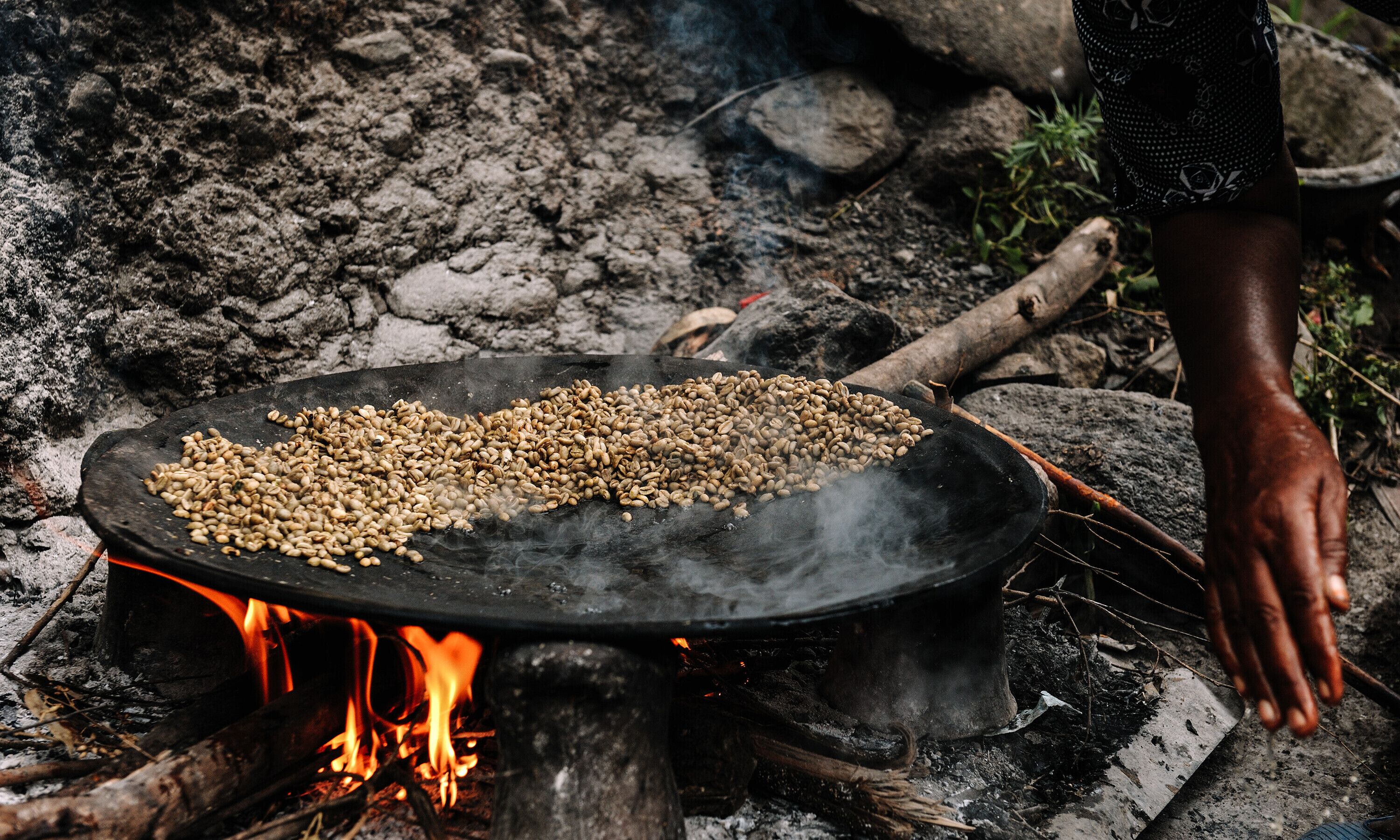 Über einem offenen Feuer liegt eine grosse, flache Pfanne, in der ein Rohstoff gebraten wird.