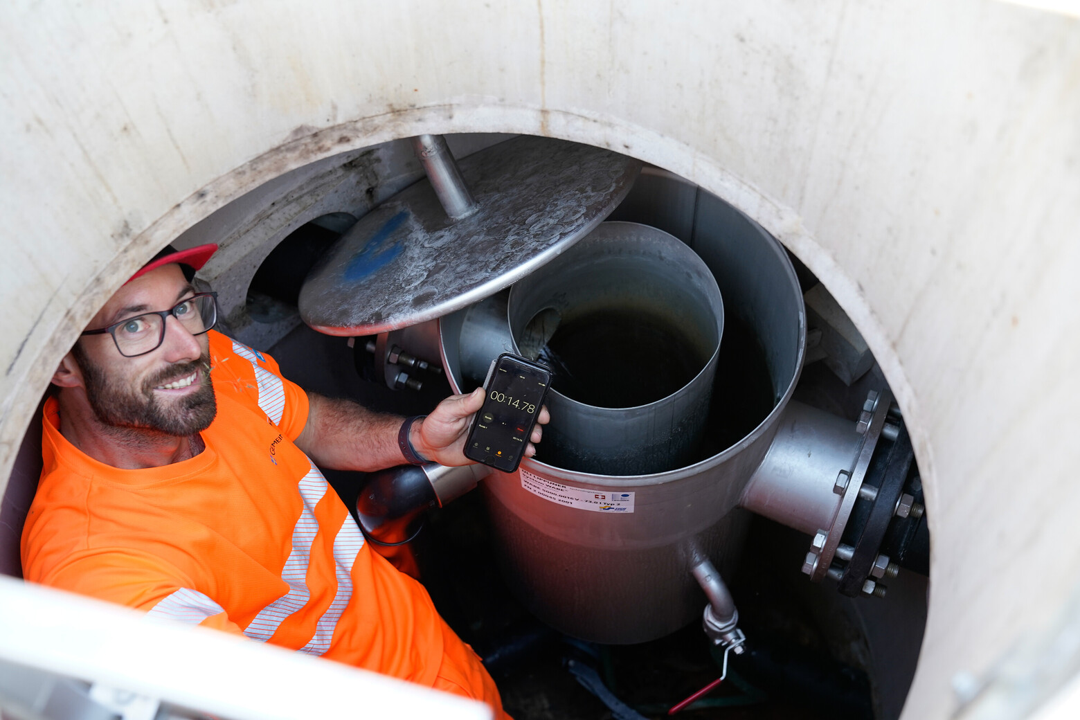 Ein Mitarbeiter der Gemeinde in orangener Schutzkleidung steht im Brunnenschacht und zeigt die gestoppte Zeit auf seinem Smartphone an. 