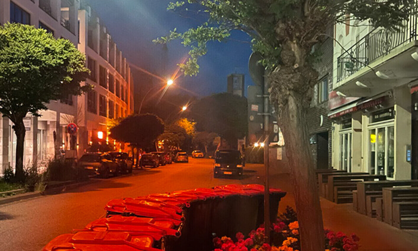 Das Bild zeigt eine Strassenflucht in einer Kleinstadt, rechts Altbauten mit einem kleinen Café, Tischen und Stühlen auf der Strasse, links Neubauten. Im Vordergrund ein blumenbepflanztes Beet, das von hohen Mülltonnen umstellt ist. Im Hintergrund sind ein paar parkierende Autos auf der Strasse und ein moderner Kirchturm zu sehen.