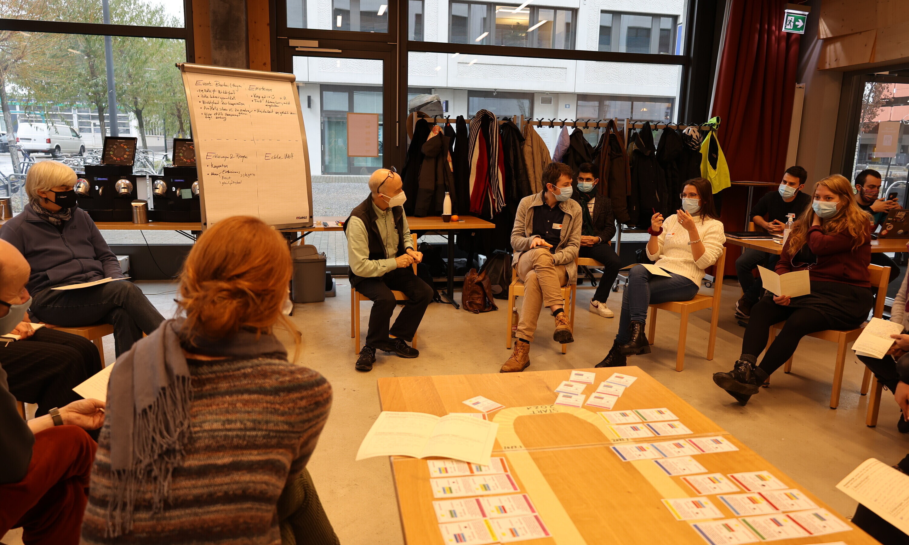 In einem Kreis aus Stühlen sitzen Männer und Frauen, die gemeinsam das Simulationsspiel „postfossilCities” spielen. In der Mitte des Kreises liegen auf dem Boden Ideen, die auf Zetteln notiert sind.