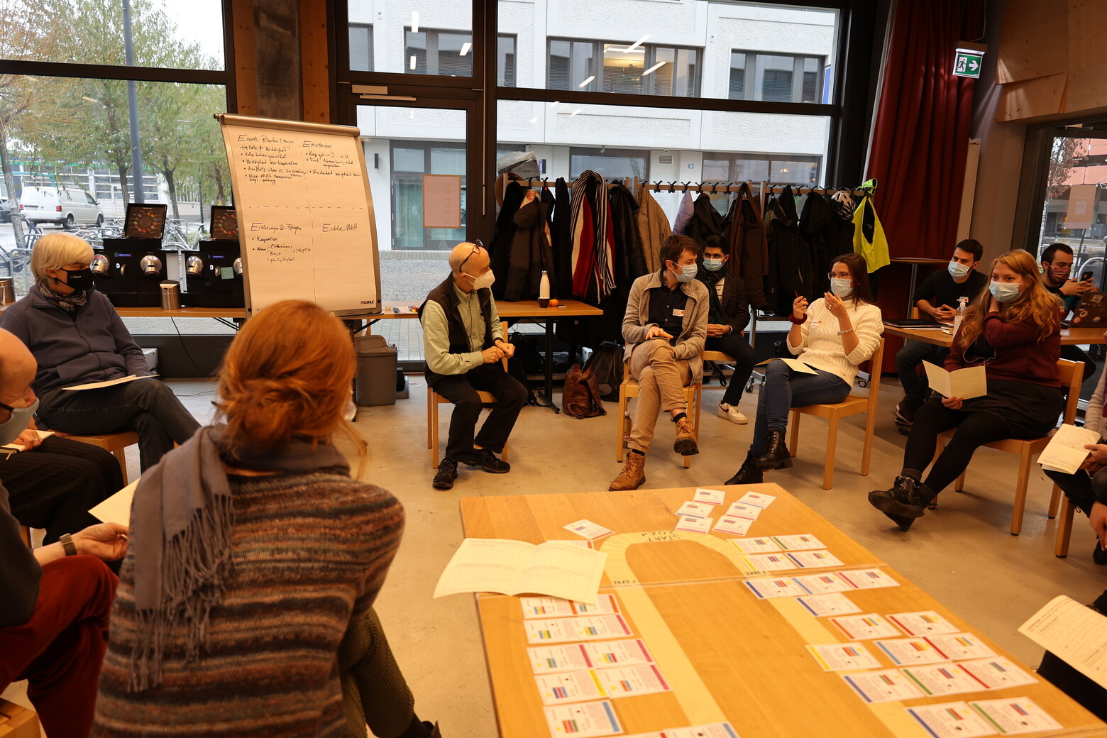 In einem Kreis aus Stühlen sitzen Männer und Frauen, die gemeinsam das Simulationsspiel „postfossilCities” spielen. In der Mitte des Kreises liegen auf dem Boden Ideen, die auf Zetteln notiert sind.