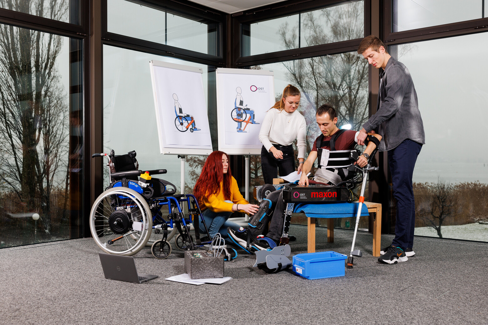 Drei Studierende rüsten einen Rollstuhlfahrer mit dem Exoskelett aus. Hinter ihnen sind die Plakate des Projekts auf einem Flipchart zu sehen.