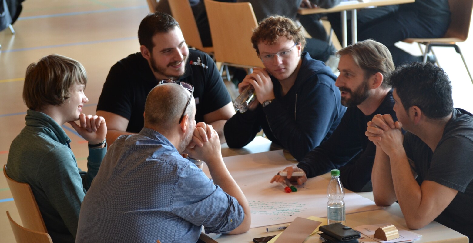 Sechs Männer sitzen an einem Tisch und brainstormen gemeinsam. Es macht ihnen sichtlich Spass, denn sie lachen.