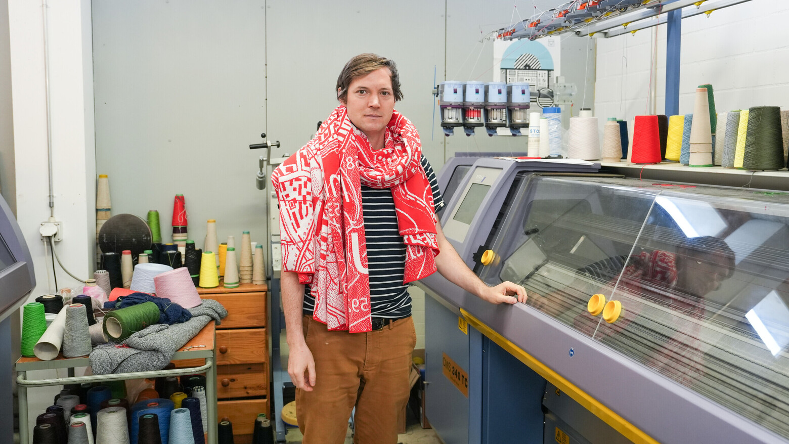 Textilunternehmer Urs Landis trägt eine Decke mit dem Stadtplan von Winterthur um den Hals und steht neben einer seiner Strickmaschinen.