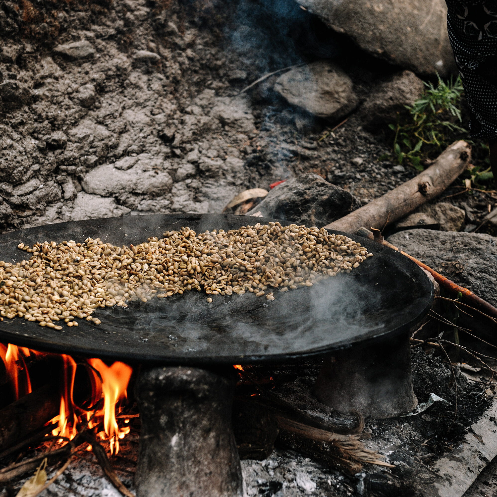 Über einem offenen Feuer liegt eine grosse, flache Pfanne, in der ein Rohstoff gebraten wird.