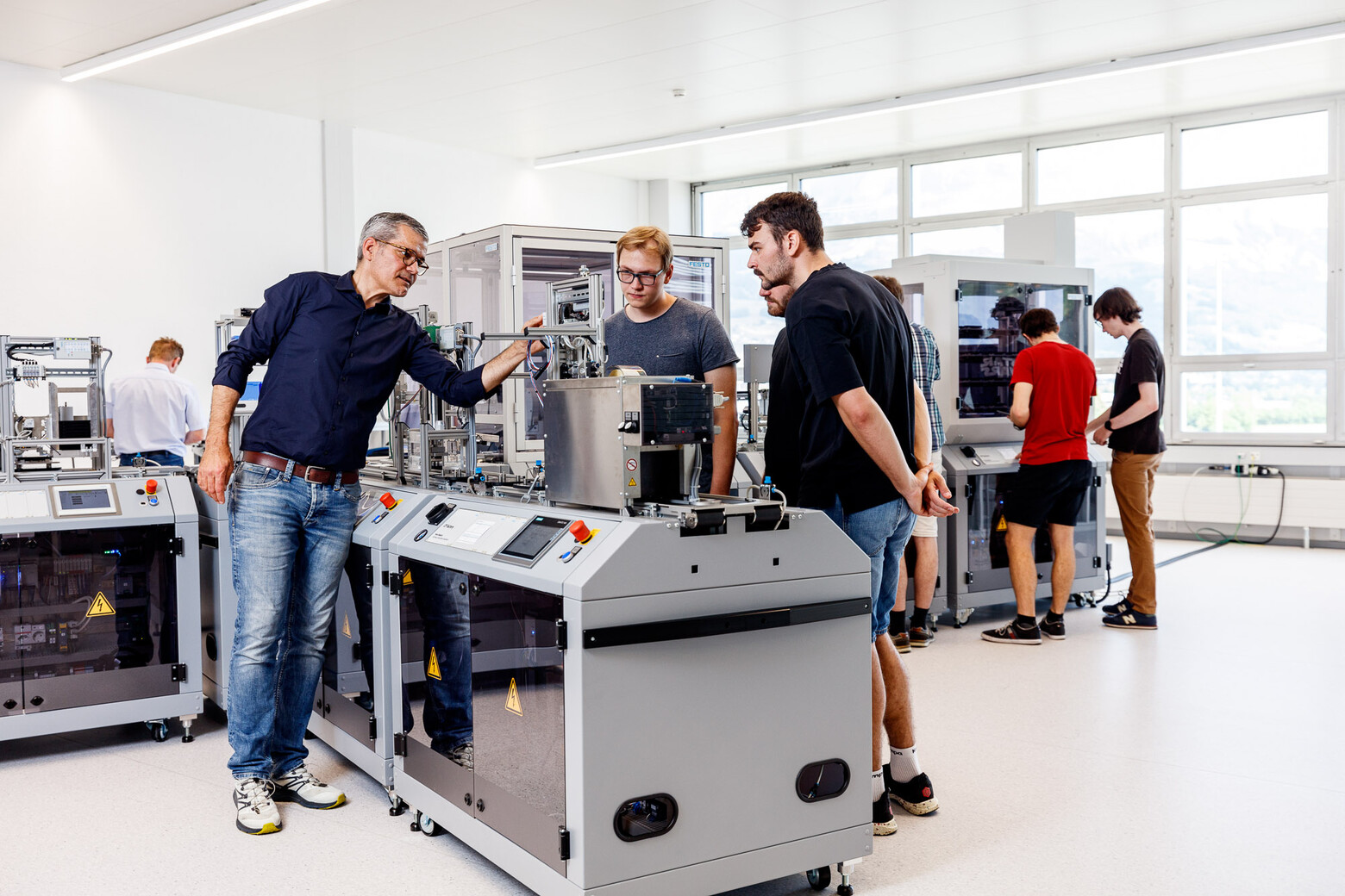  In der Produktion sind interessierte Gesichter zu sehen. Ein Mann erklärt den Studierenden, die um die Maschine stehen, den Ablauf.