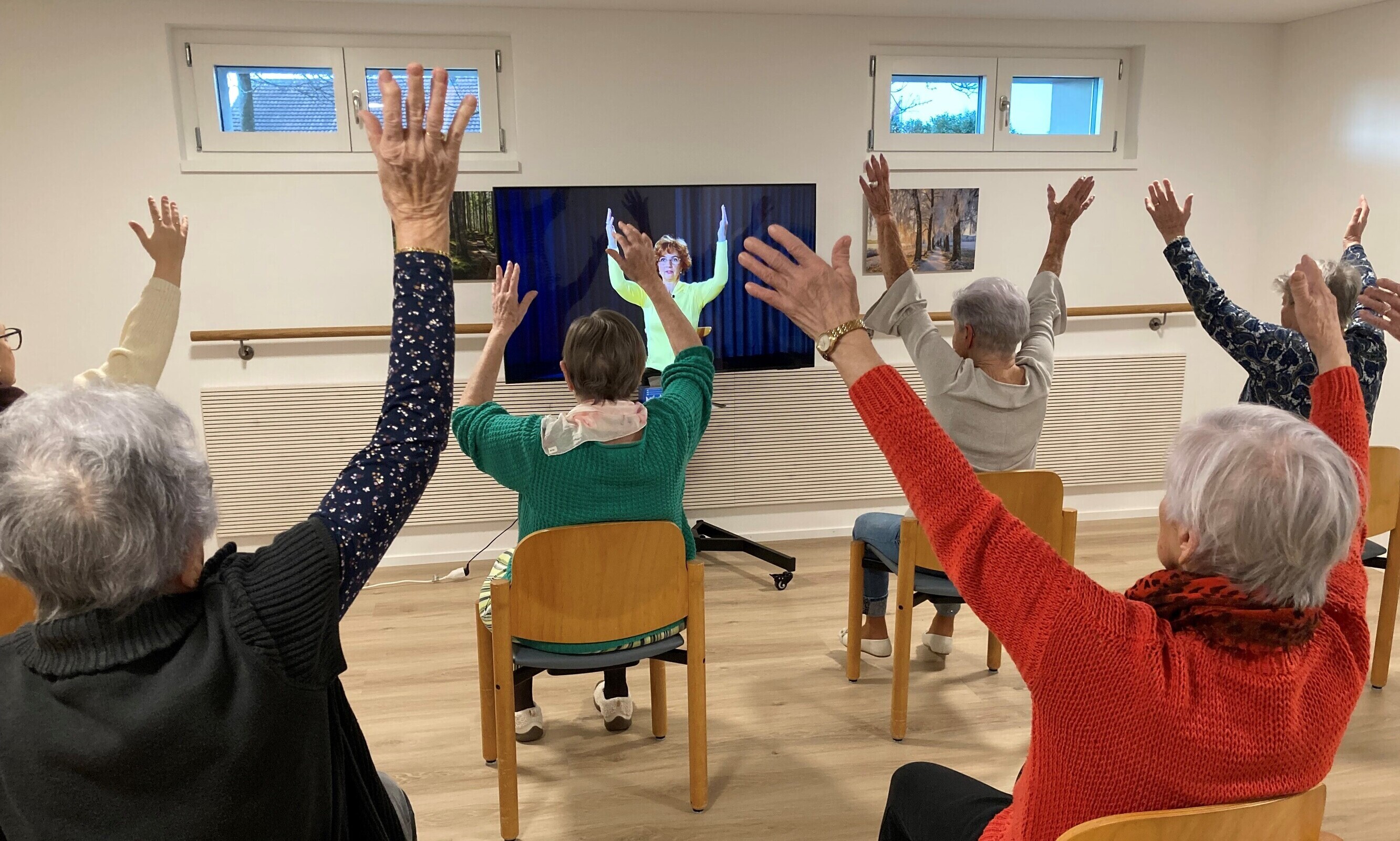 Fünf ältere Damen werden von hinten fotografiert, wie sie auf ihren Stühlen die Bewegungen aus dem Fernseher nachturnen.