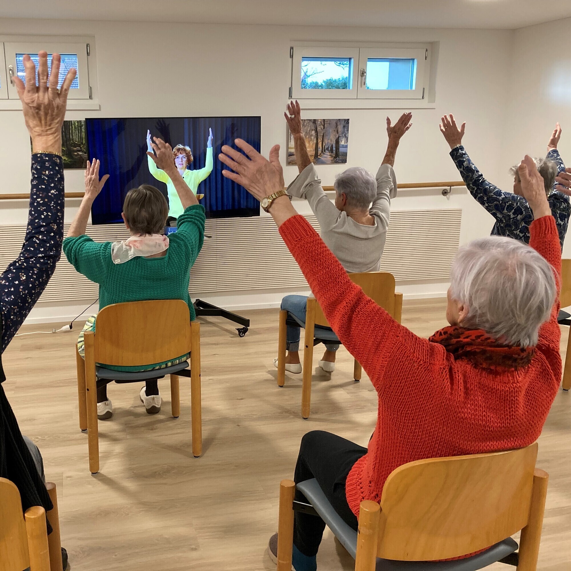Fünf ältere Damen werden von hinten fotografiert, wie sie auf ihren Stühlen die Bewegungen aus dem Fernseher nachturnen.