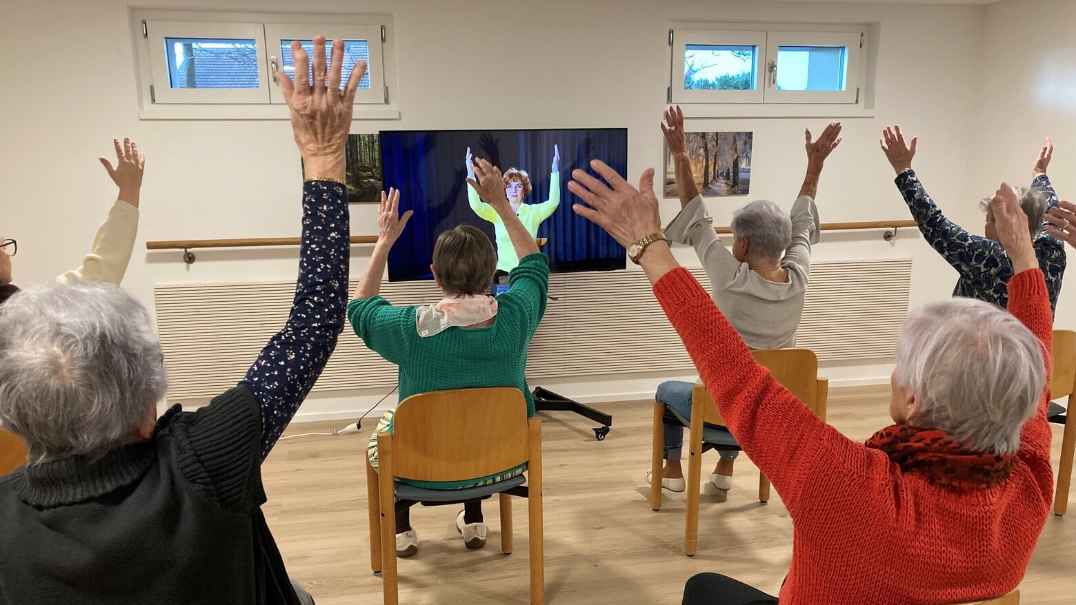 Fünf ältere Damen werden von hinten fotografiert, wie sie auf ihren Stühlen die Bewegungen aus dem Fernseher nachturnen.