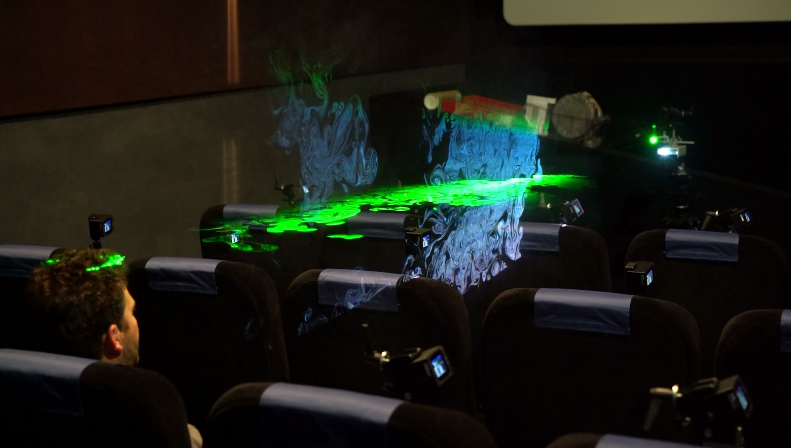 Ein Mann sitzt auf einem Kinosessel, während Laser- und Lichtstrahlen das Unsichtbare sichtbar machen.