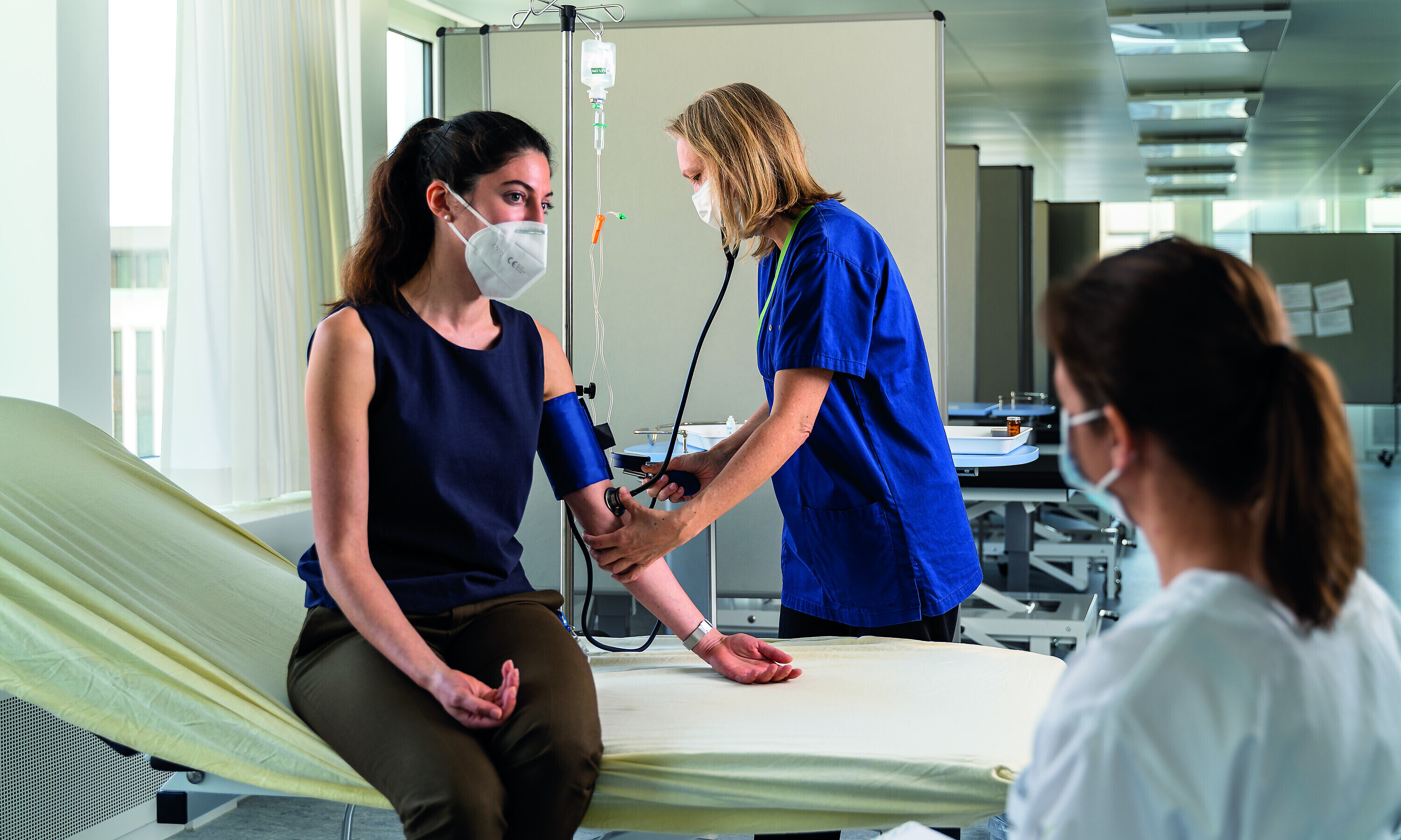 Während eine Ärztin mit der Patientin spricht, wird ihr auf dem Krankenbett der Puls gemessen. Eine Pflegeassistentin hält den blauen Verband.