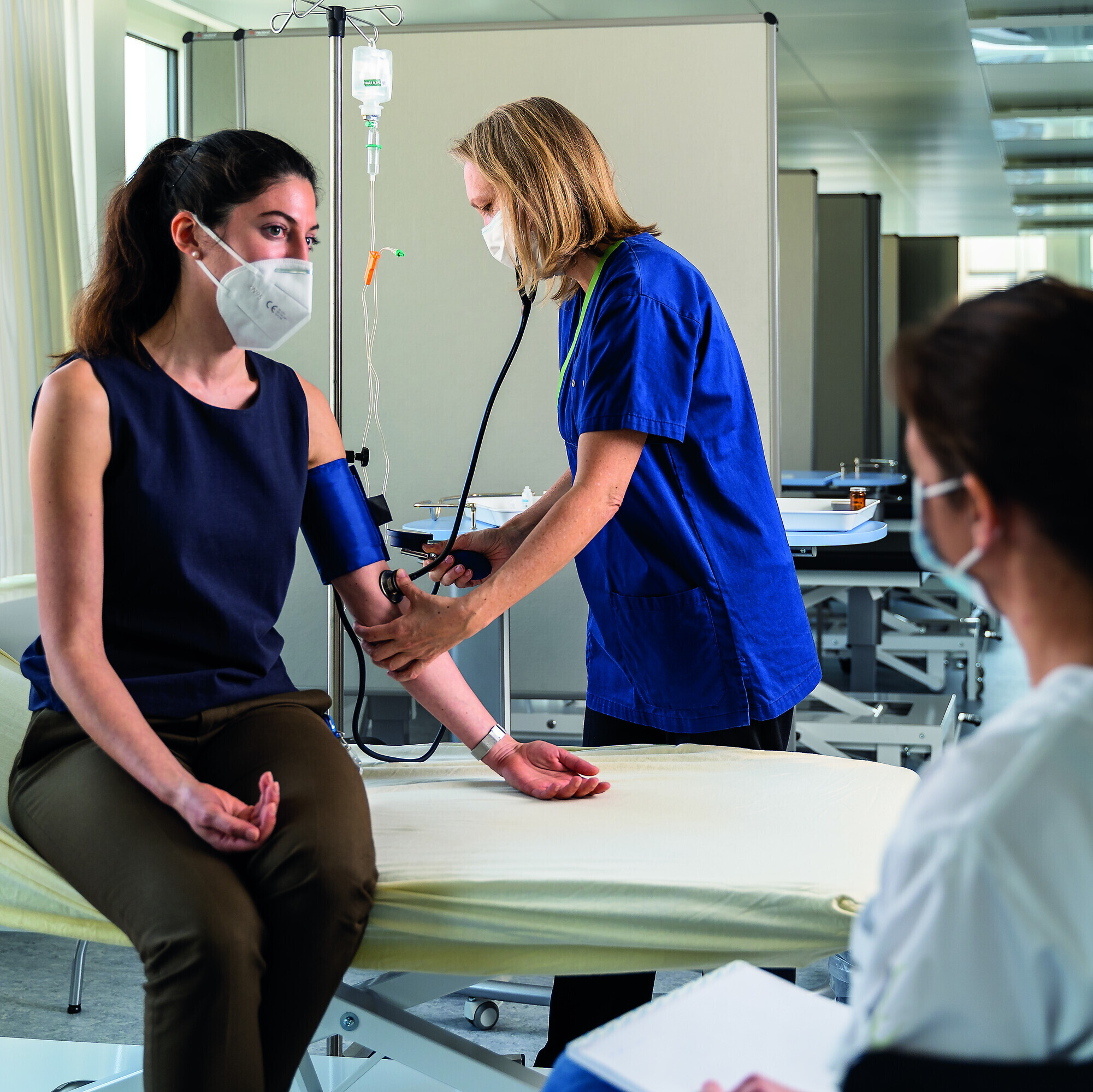 Während eine Ärztin mit der Patientin spricht, wird ihr auf dem Krankenbett der Puls gemessen. Eine Pflegeassistentin hält den blauen Verband.
