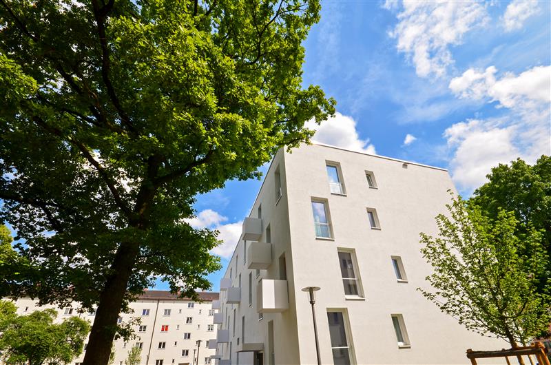 Vor dem Mehrfamilienhaus stehen Bäume mit sattgrünen Blättern.